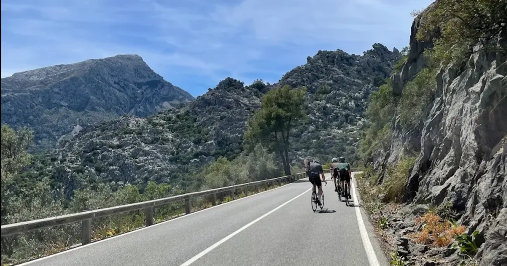 Three riders on a wide, smooth road under a light blue sky, guardrail on the left and cut rock on the right.