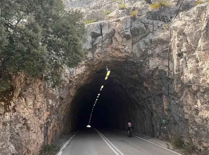 Boca de túnel oscura abierta en la roca caliza desnuda, un ciclista entrando bajo una hilera de luces amarillas.
