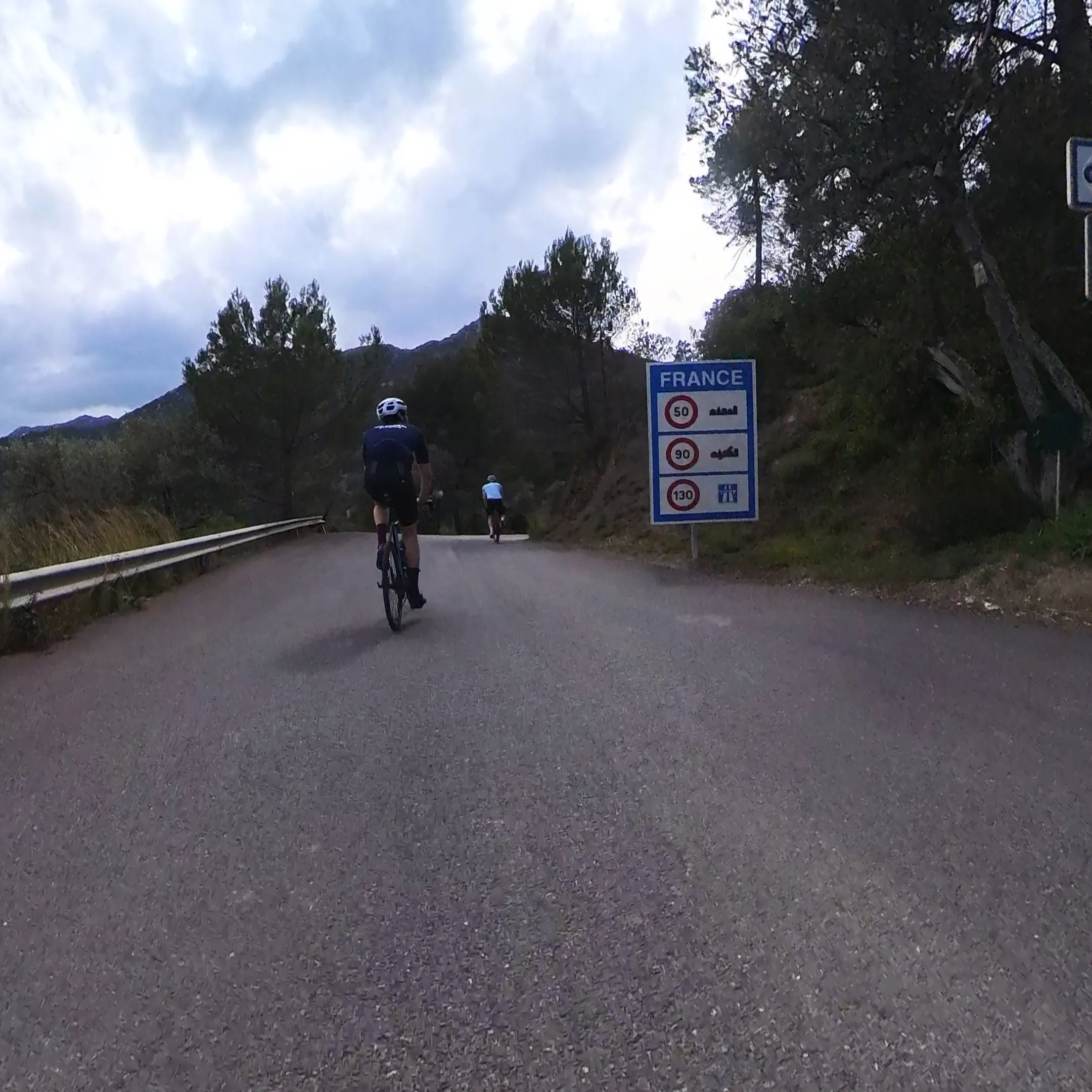 Radfahrer passieren das FRANCE-Schild am Col de Vescavo, schmaler Asphalt und Leitplanke über Olivetta San Michele.