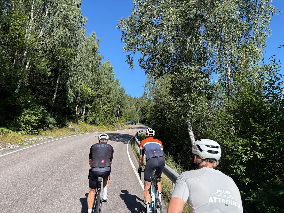 Tres ciclistas en el tramo medio de Rundvollbakken, con bosque a ambos lados