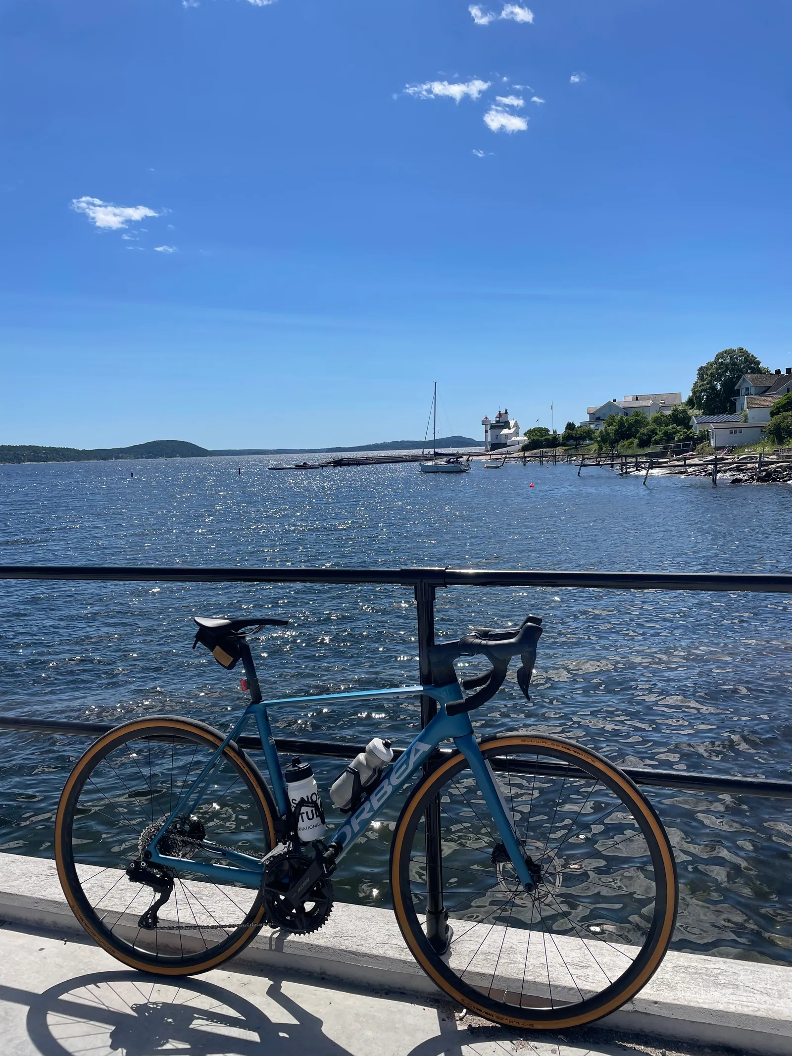 Orbea azul apoyada en el muelle, con un velero y el faro de Filtvet al fondo.