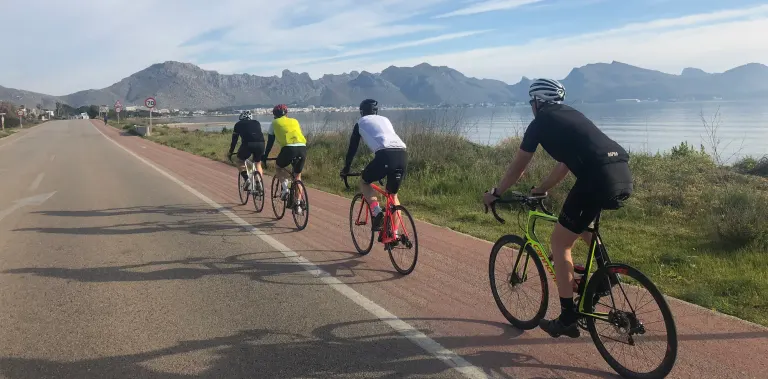 Cyclists on the flat coastal road in Bay of Alcúdia, with the Cap de Formentor mountain range visible across the water, Mallorca.