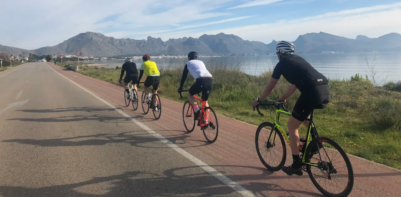 Cyclists on the flat coastal road in Bay of Alcúdia, with the Cap de Formentor mountain range visible across the water, Mallorca.