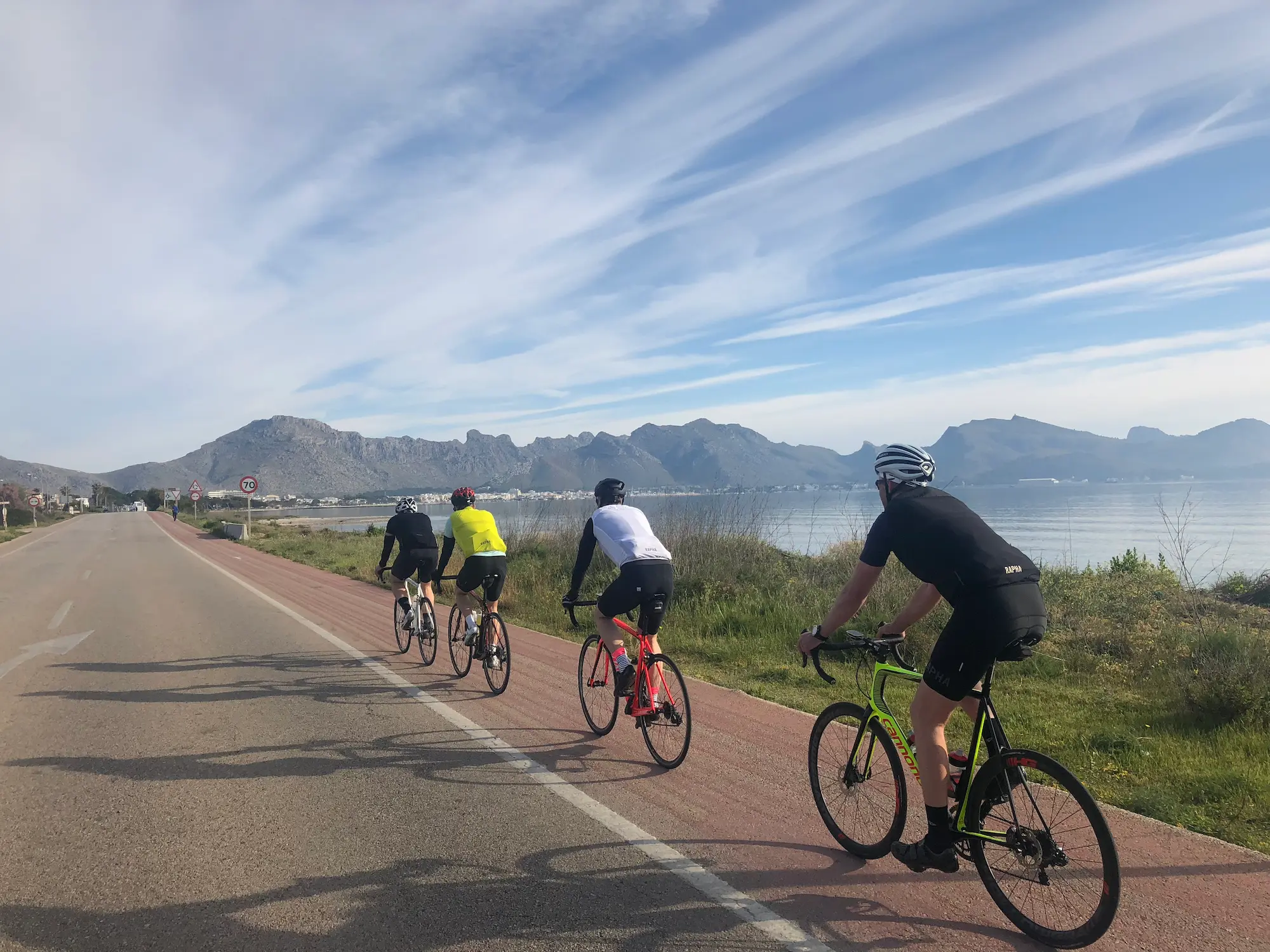 Cycling along the coastal road from Can Picafort toward Alcúdia, with Cap de Formentor in the distance, blue sky over the bay.
