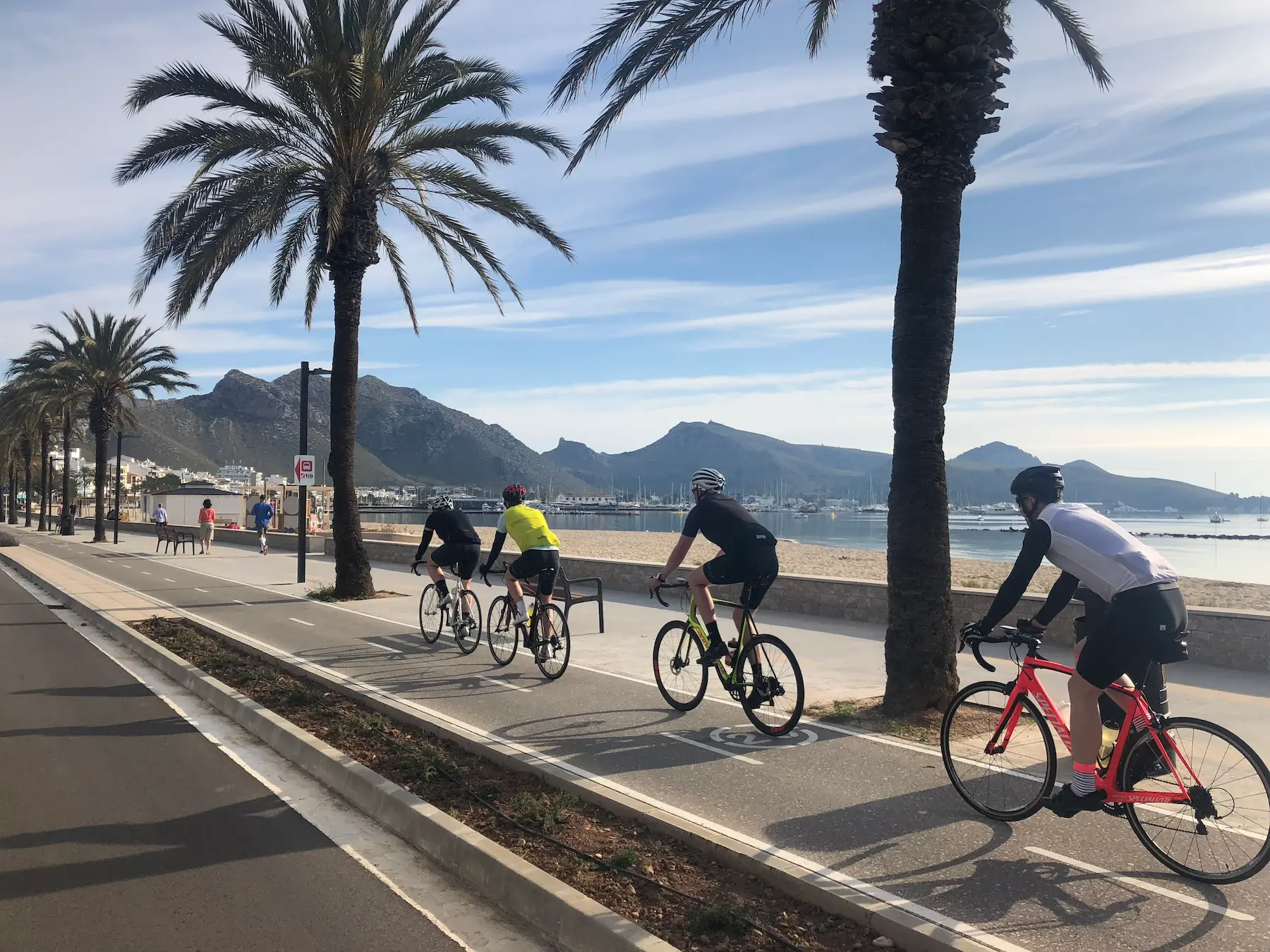 Road cyclists in Bay of Alcúdia on a flat Mallorca rest-day ride, Cap de Formentor mountains across the sea in the background.
