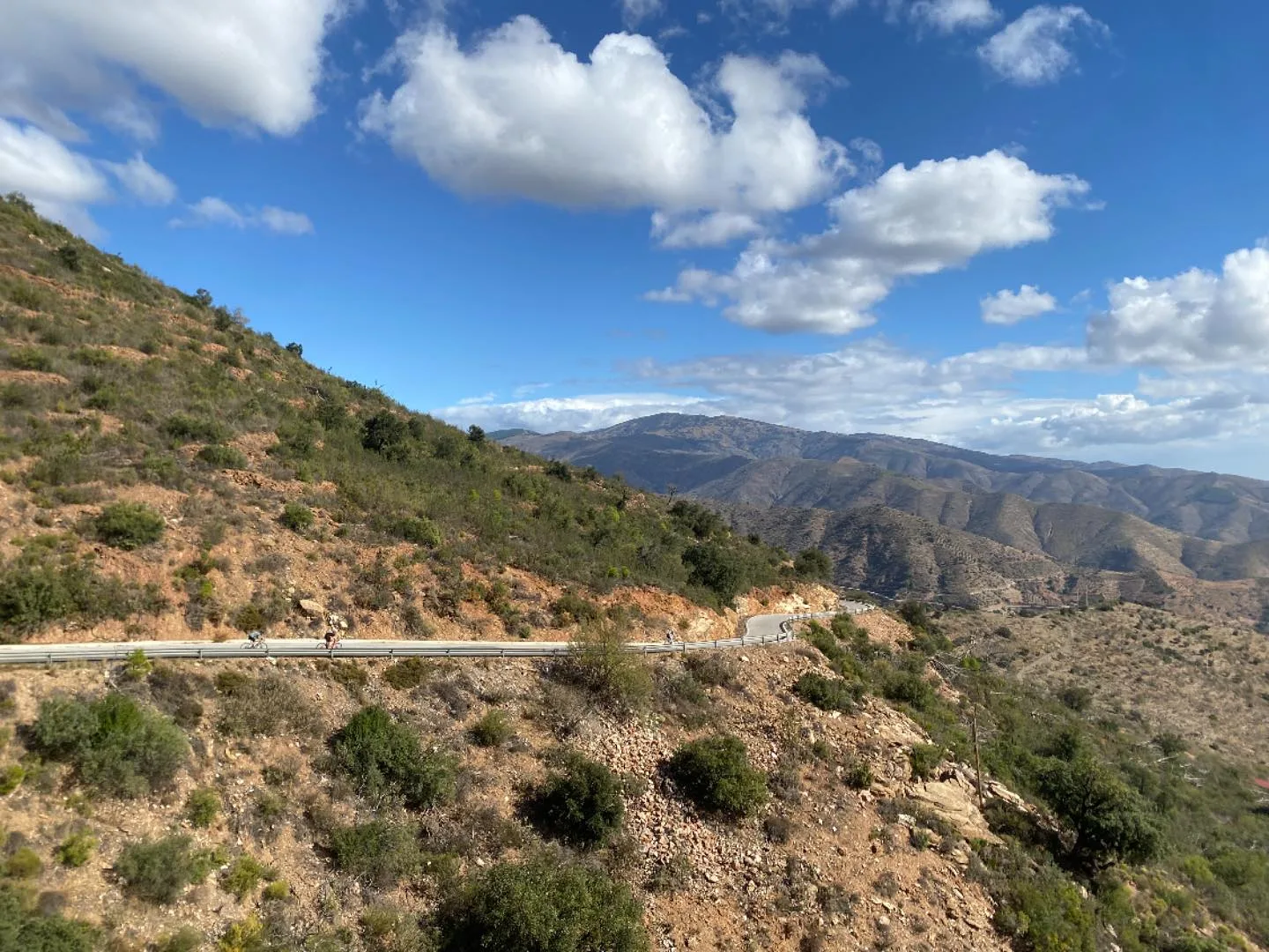 Tres ciclistas en carretera con quitamiedos, ladera rojiza y crestas al fondo